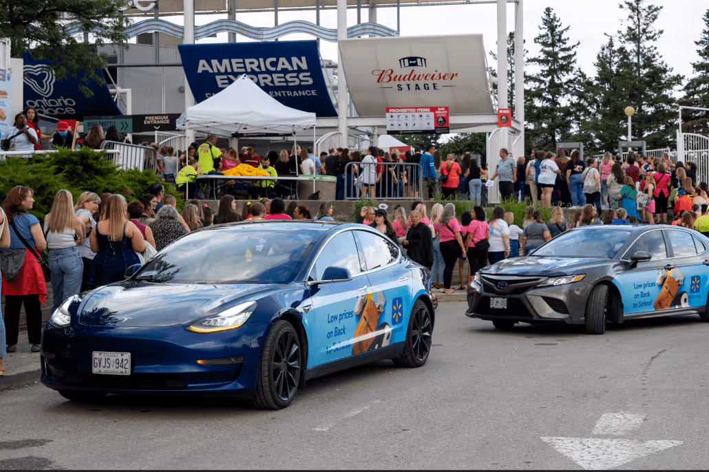 Two cars with Walmart advertisements for low back-to-school prices parked near a crowd outside the American Express entrance at Budweiser Stage.