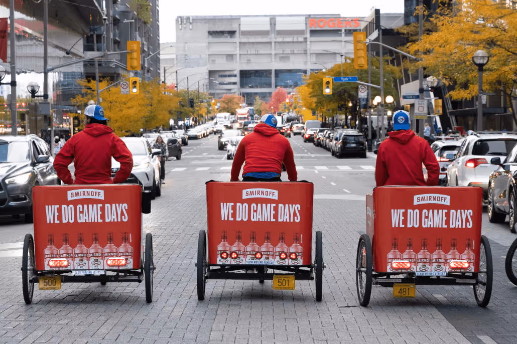 Three people in red hoodies riding pedicabs with Smirnoff ads reading 'We Do Game Days' on a city street lined with autumn trees.