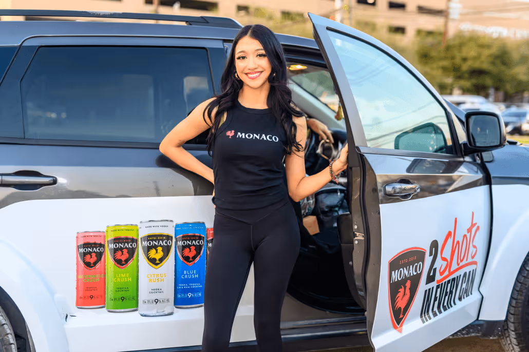 Smiling woman in black outfit standing next to an open door of a black and white vehicle with Monaco drink brand logos and cans displayed on the side.