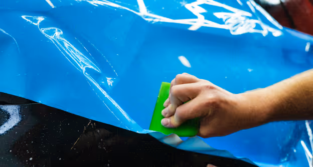 Hand smoothing blue vinyl wrap on a black car surface using a green squeegee tool.