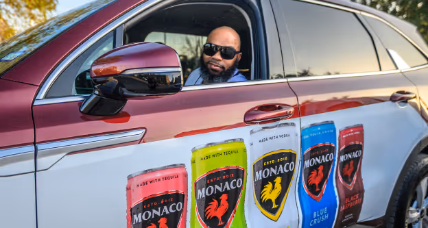 Man wearing sunglasses sitting inside a red car with Monaco flavored beverage can designs on the door panel.
