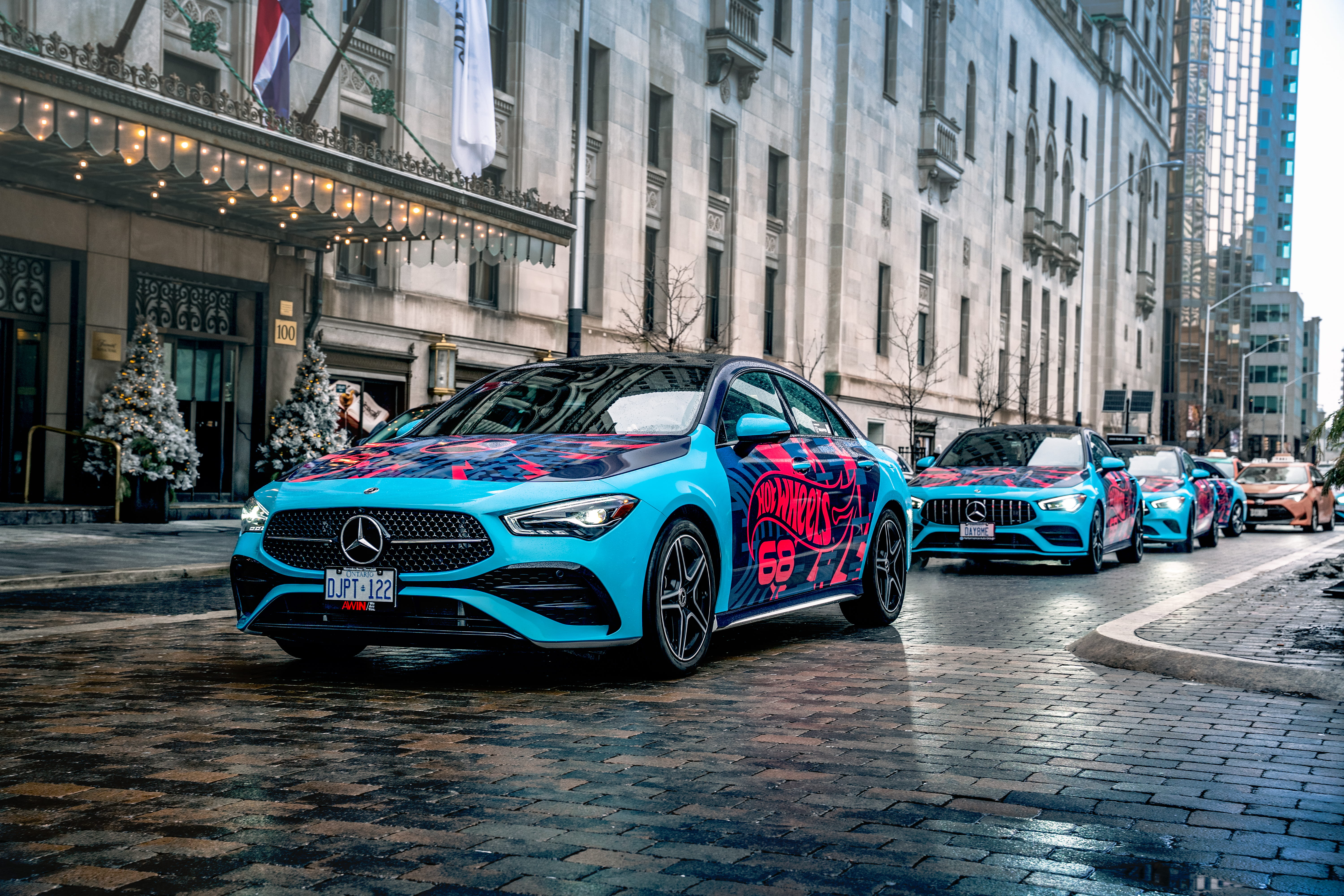 Line of blue Mercedes-Benz cars with Hot Wheels branding parked on a cobblestone street in front of a building with festive decorations.