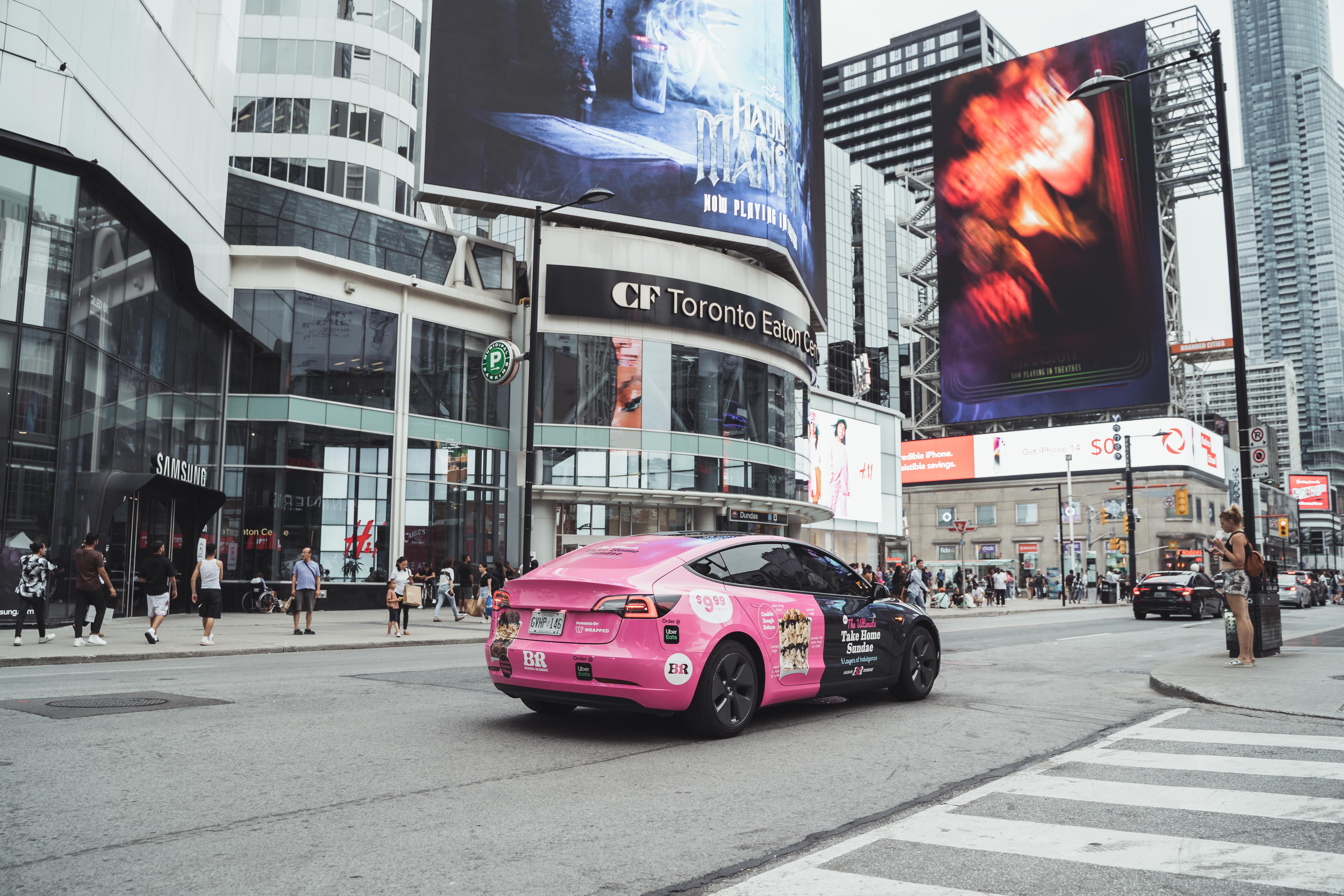 Pink and black car with Baskin Robbins ice cream ad driving near CF Toronto Eaton Centre with pedestrians and tall buildings around.