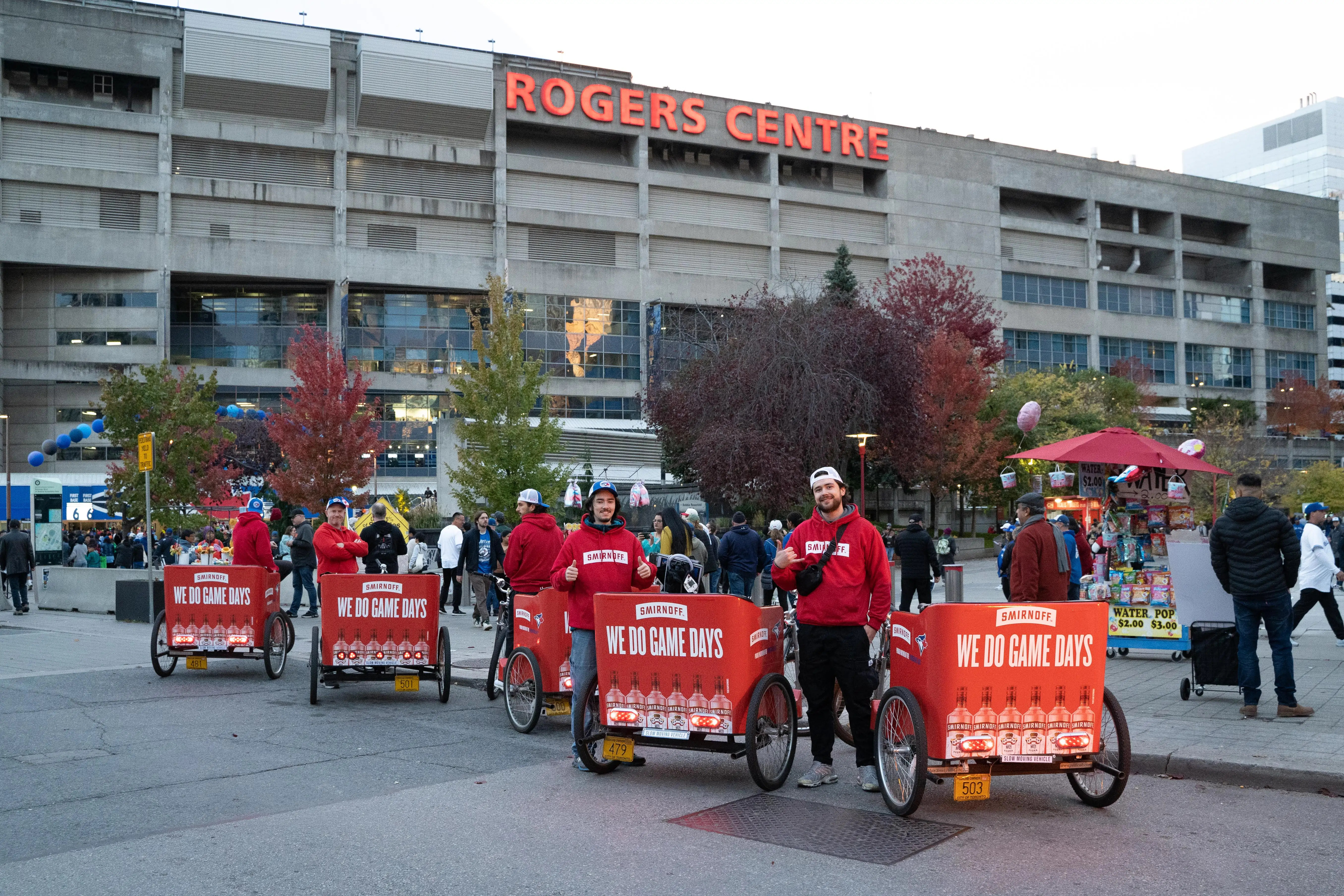 Four men in red Smirnoff hoodies with red bike trailers that say 'WE DO GAME DAYS' outside Rogers Centre with crowd and trees in the background.