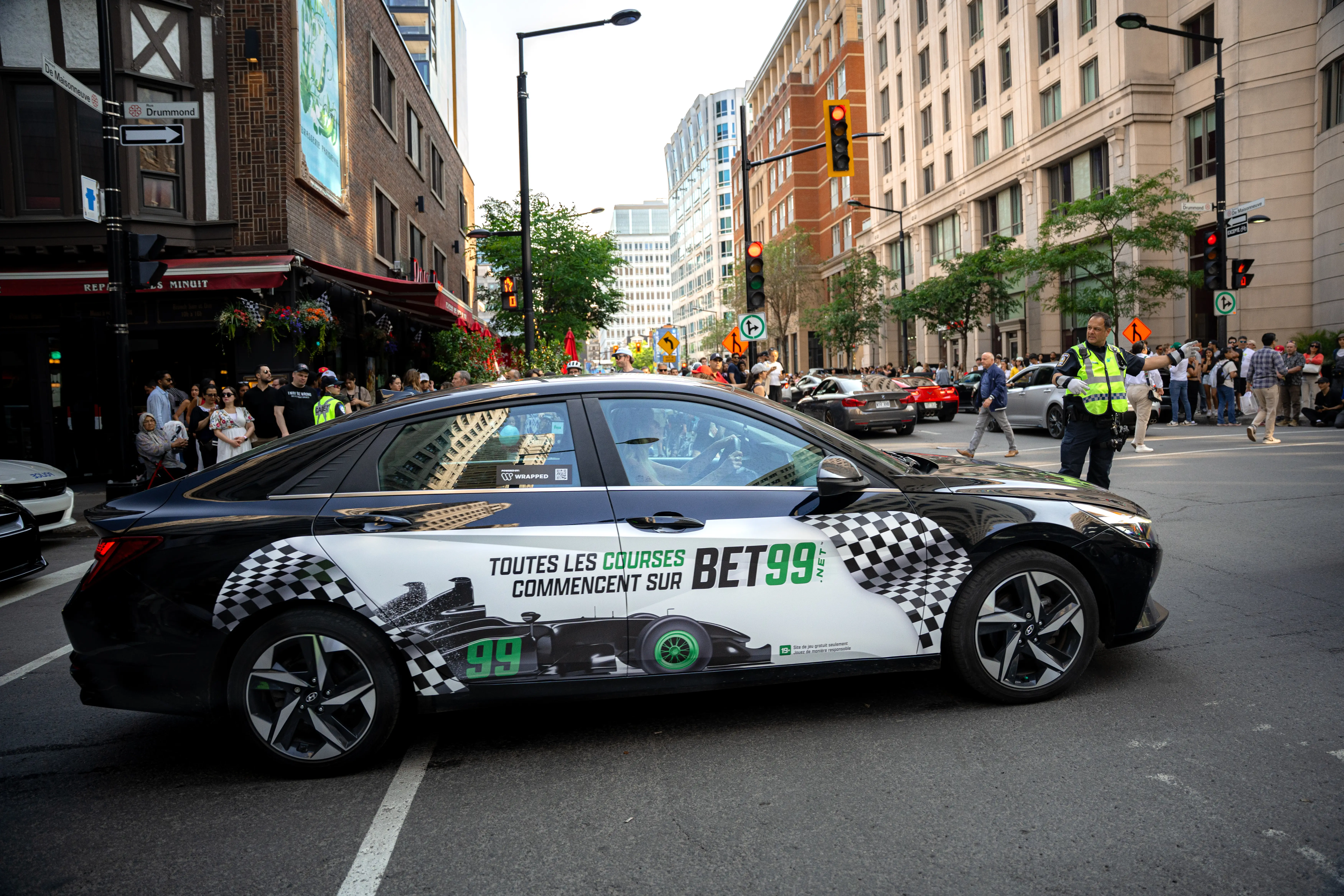 Black and white car with checkered racing design and text promoting BET99 on a city street with crowd and traffic officer.