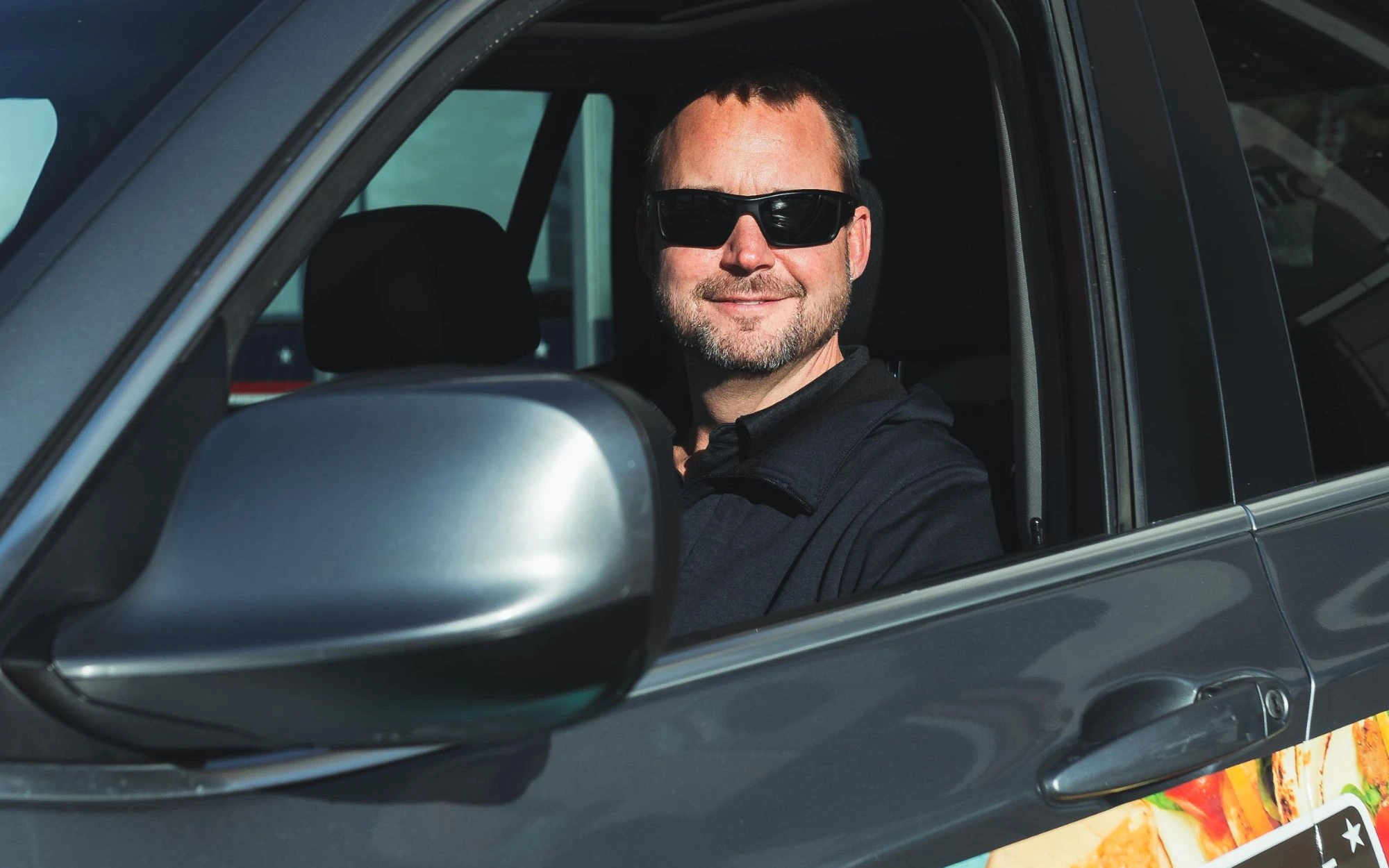 Man wearing sunglasses smiling while sitting in the driver's seat of a black vehicle.