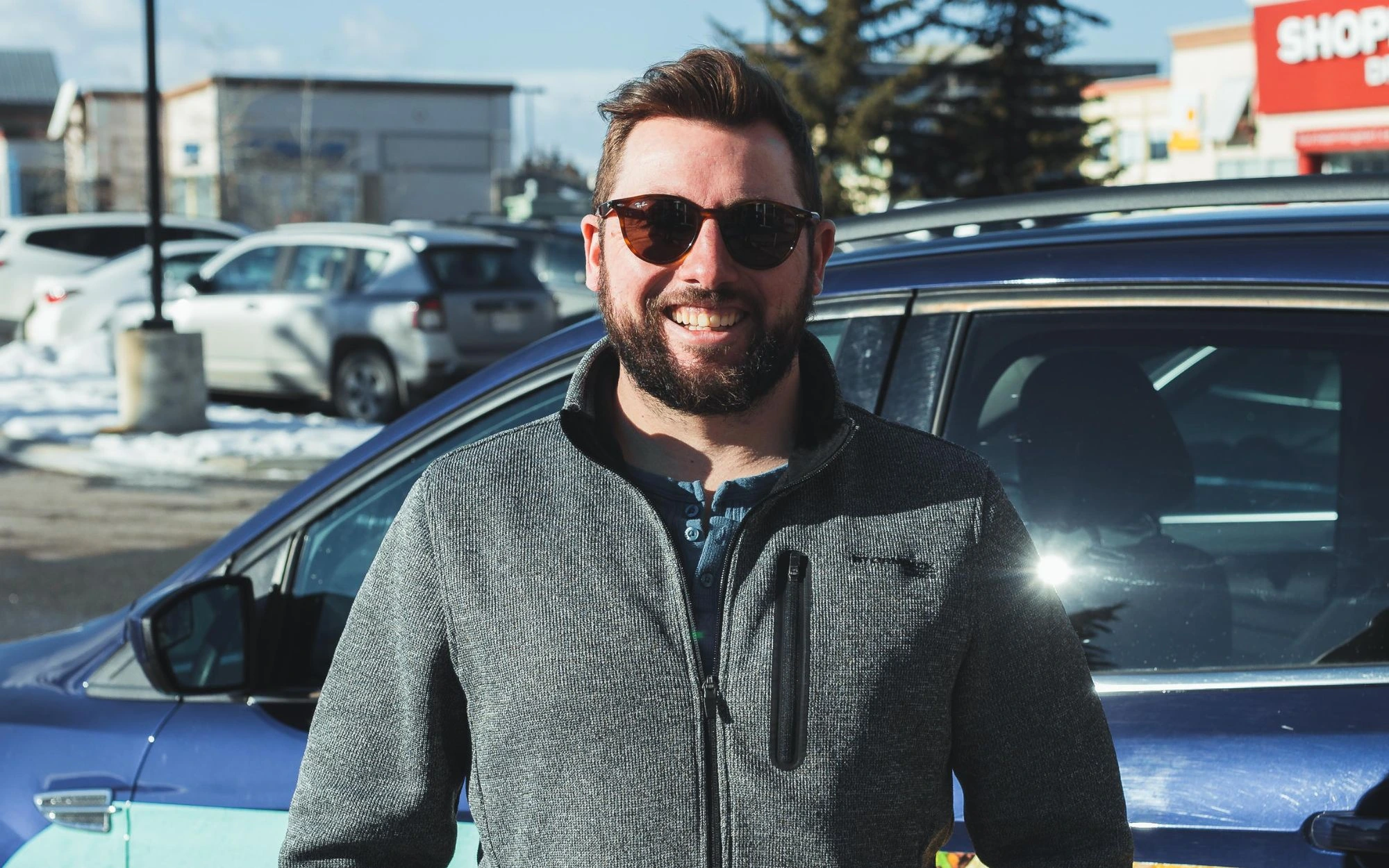 Smiling man with beard and sunglasses standing outdoors near a blue car in a snowy parking lot.