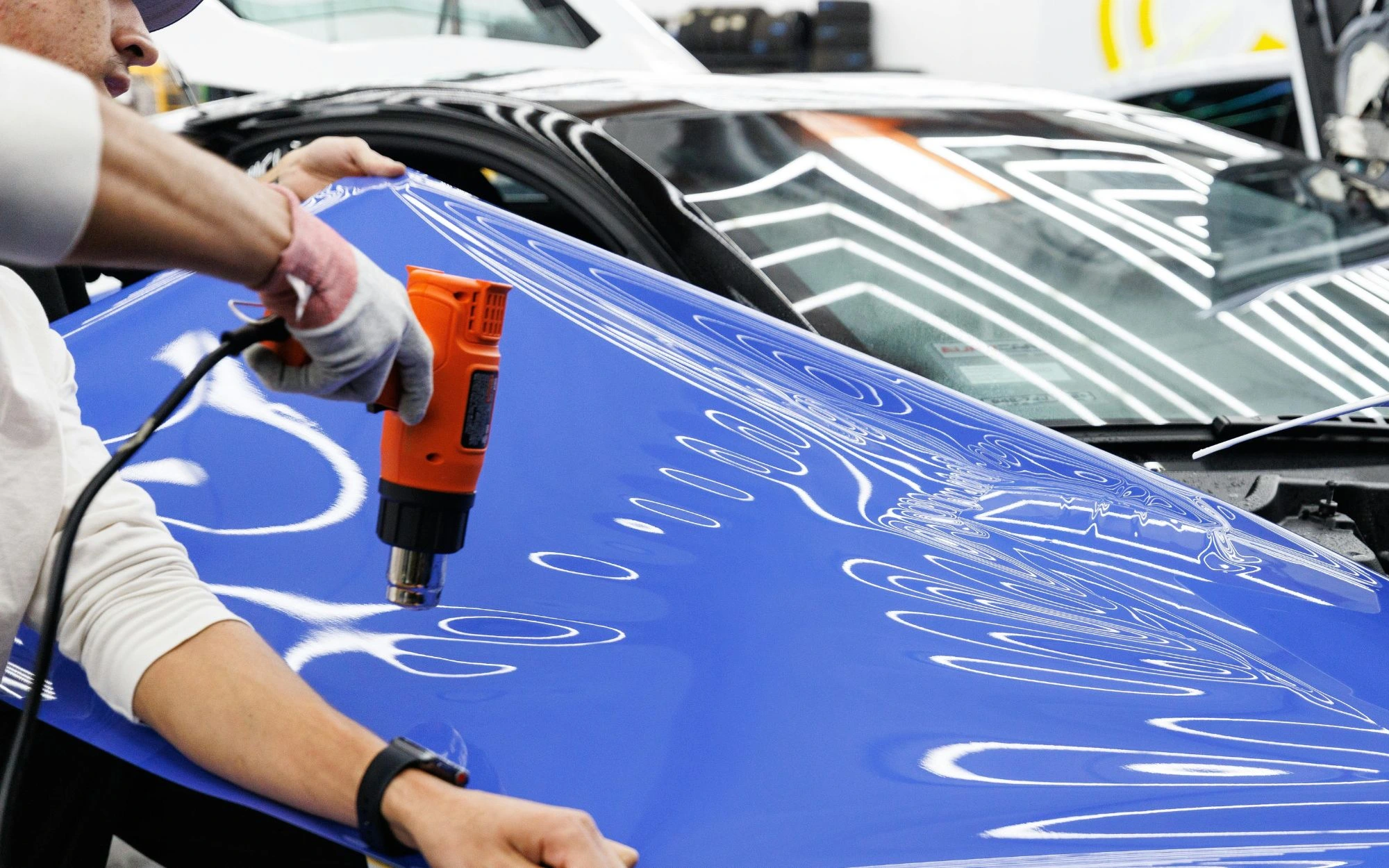 Person applying a blue vinyl wrap to a car hood using an orange heat gun.