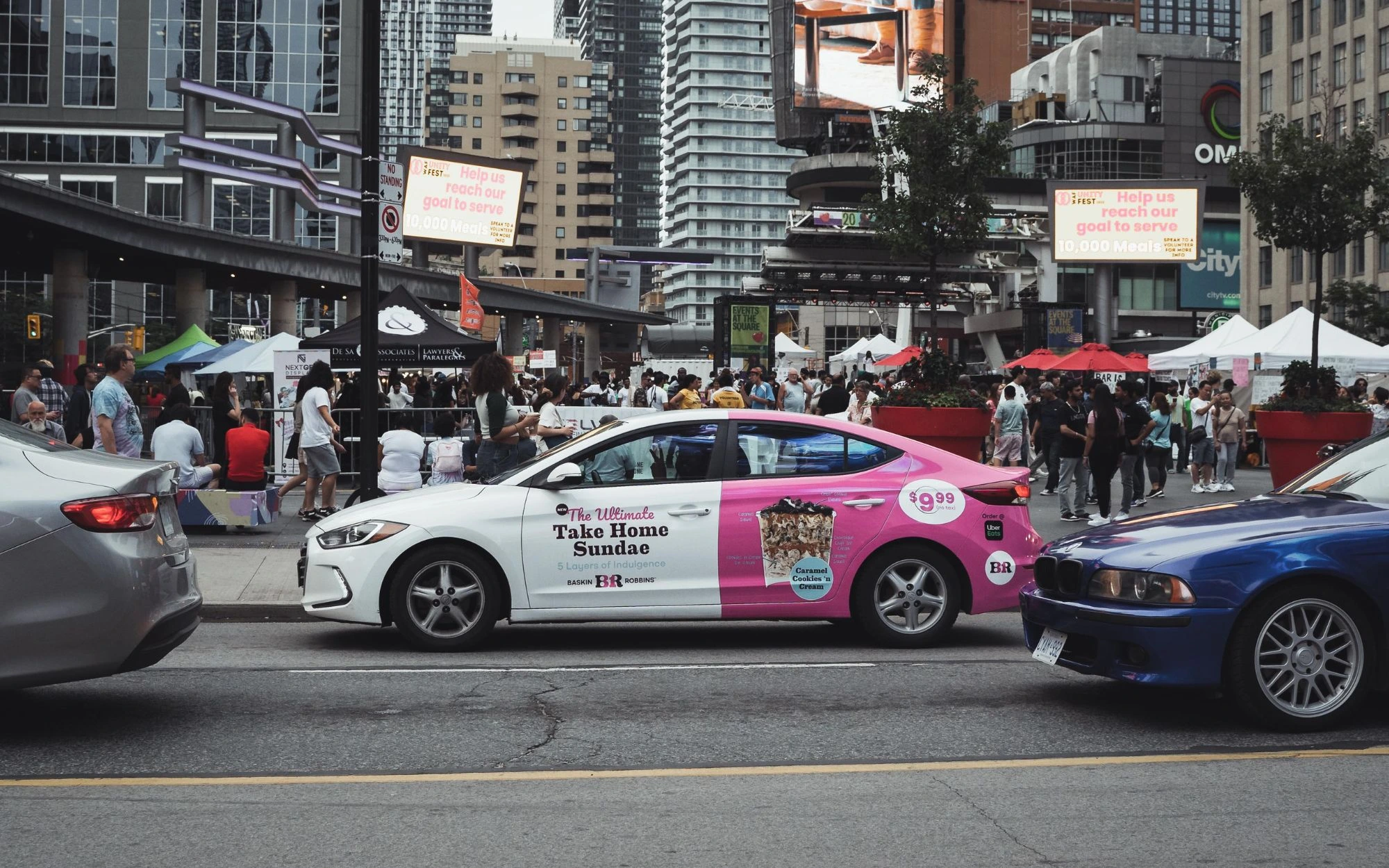 White and pink car with Baskin Robbins advertisement for The Ultimate Take Home Sundae parked on a city street with a crowd and tents in the background.