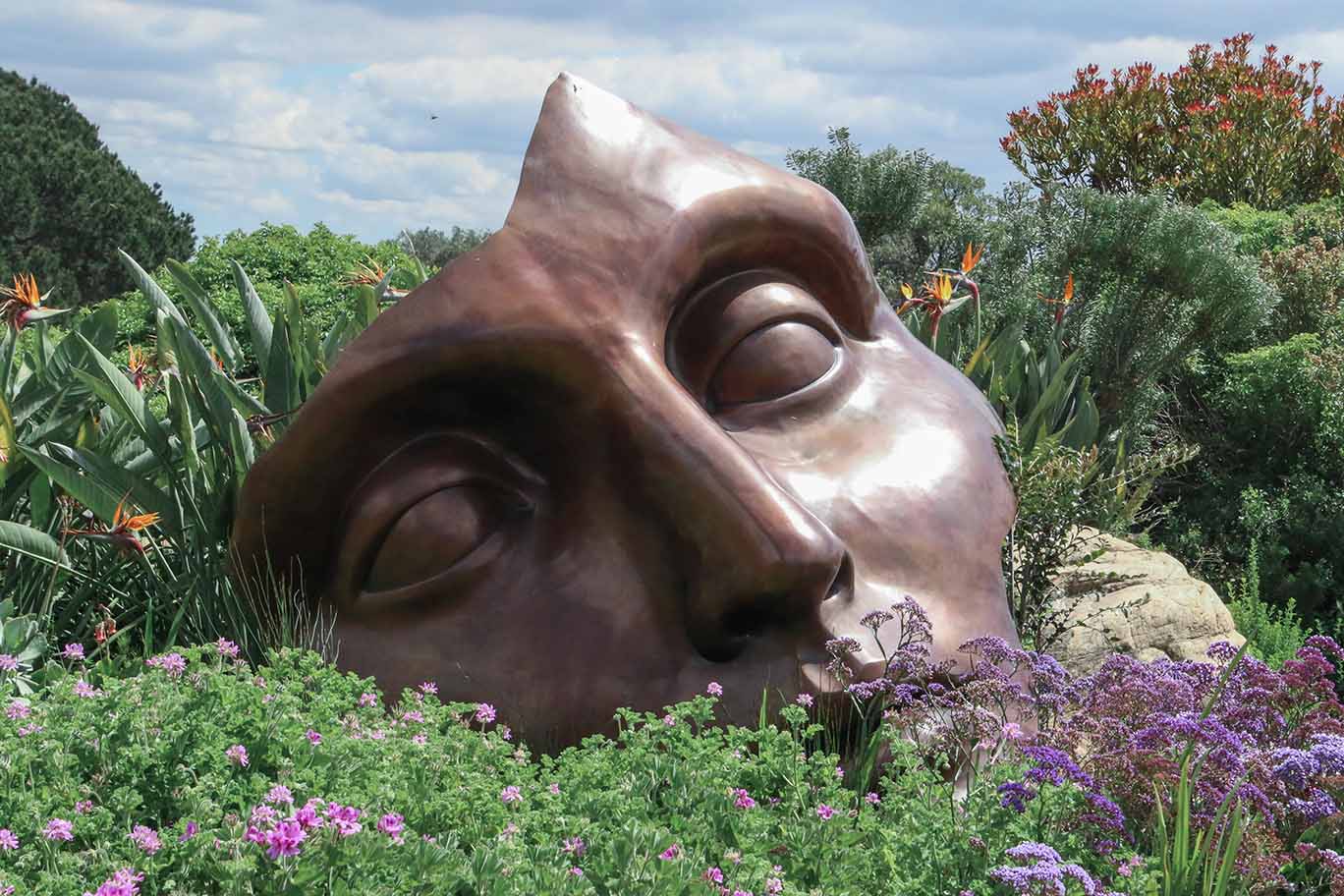 Large bronze sculpture of a human face partially buried among green plants and colorful flowers in an outdoor garden.