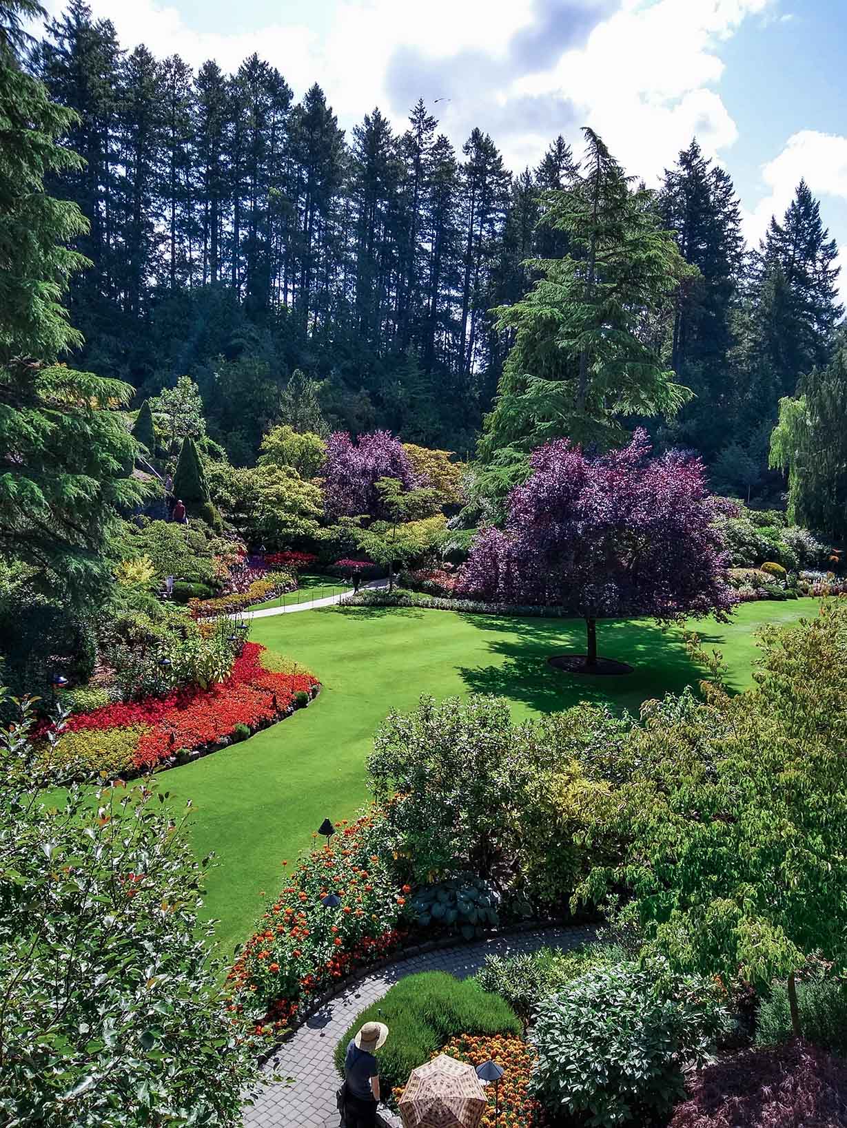 A vibrant garden with colorful flower beds, green lawn, tall trees, and two people walking on a stone path.