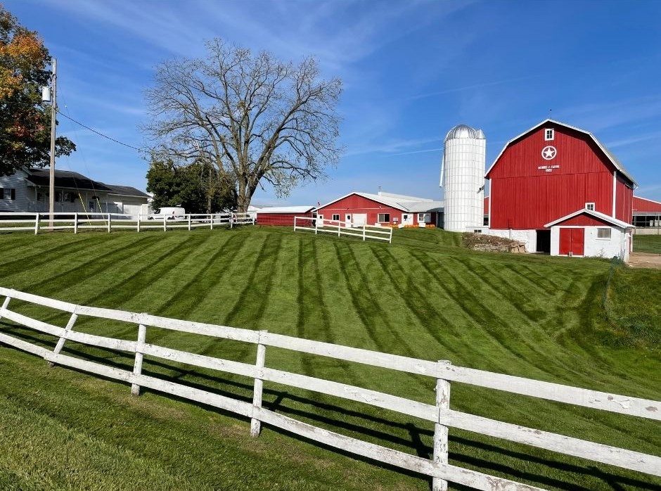 Green lawn with mowing stripes in front of a white fence, large bare tree, white farmhouse, and red barn with silo under blue sky.
