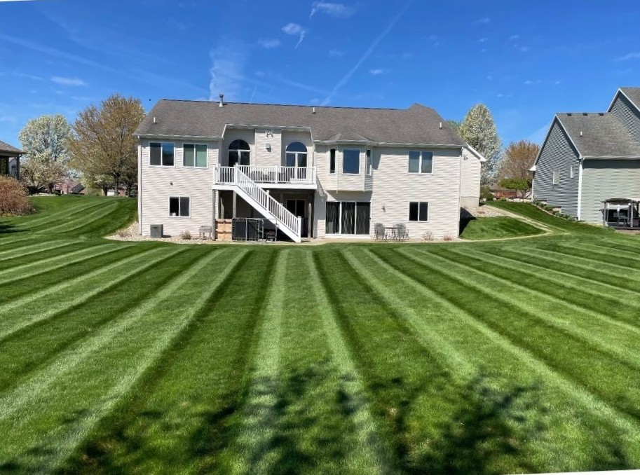 Back view of a two-story house with white siding and a large, evenly striped green lawn under a blue sky.