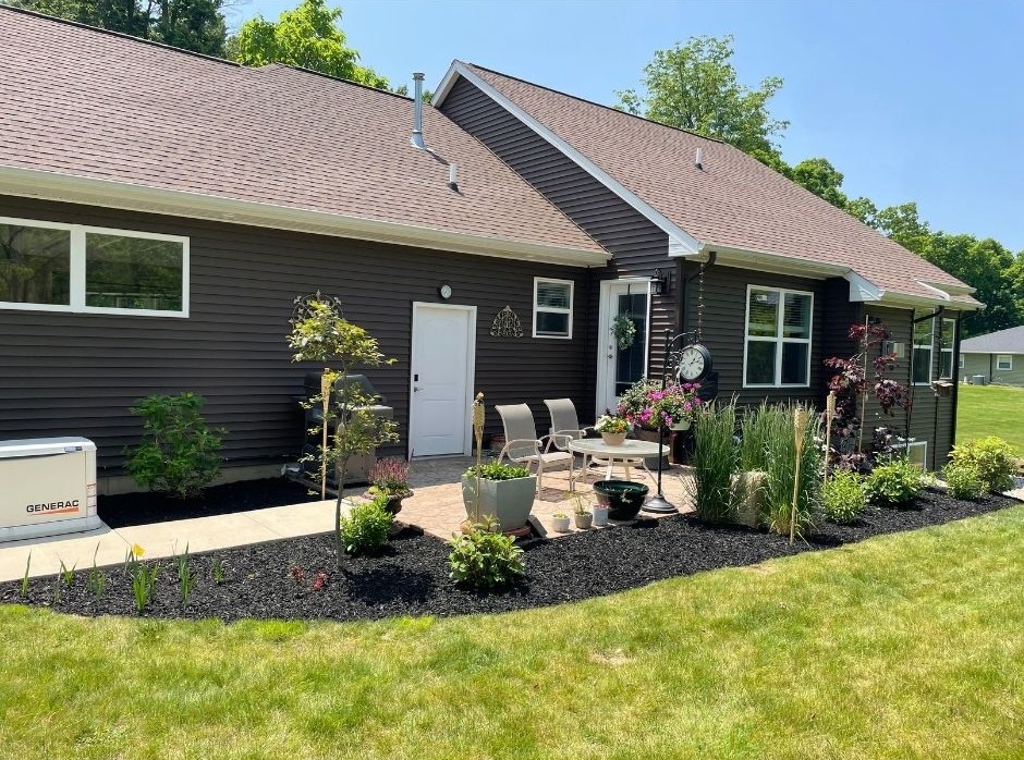 Backyard patio with chairs, a round table, potted plants, and a garden clock next to a dark brown house with light tan roof shingles.