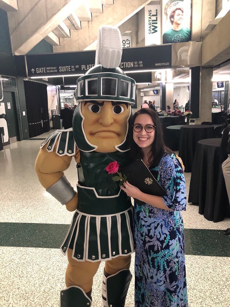 A smiling woman in glasses and a blue patterned dress stands next to a Spartan mascot holding a pink rose inside a stadium concourse.