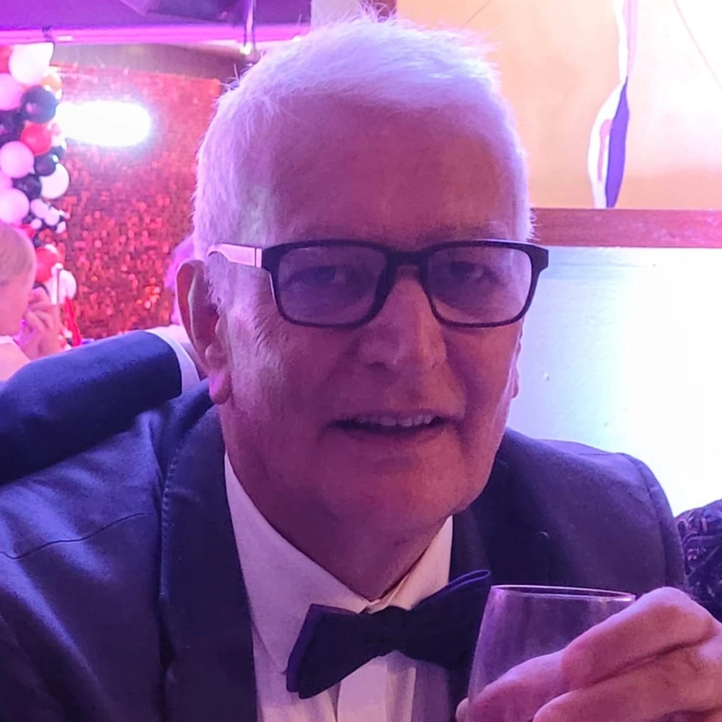 Elderly man wearing glasses, a black tuxedo, and a bow tie, holding a glass in a celebratory setting with balloons in the background.