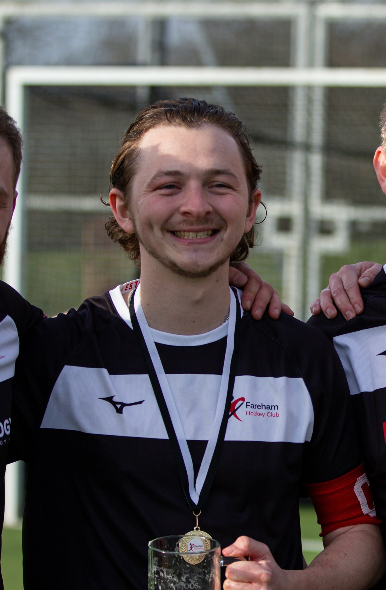 Smiling young man wearing a Fareham Hockey Club jersey with a medal around his neck, holding a glass trophy on a sports field.