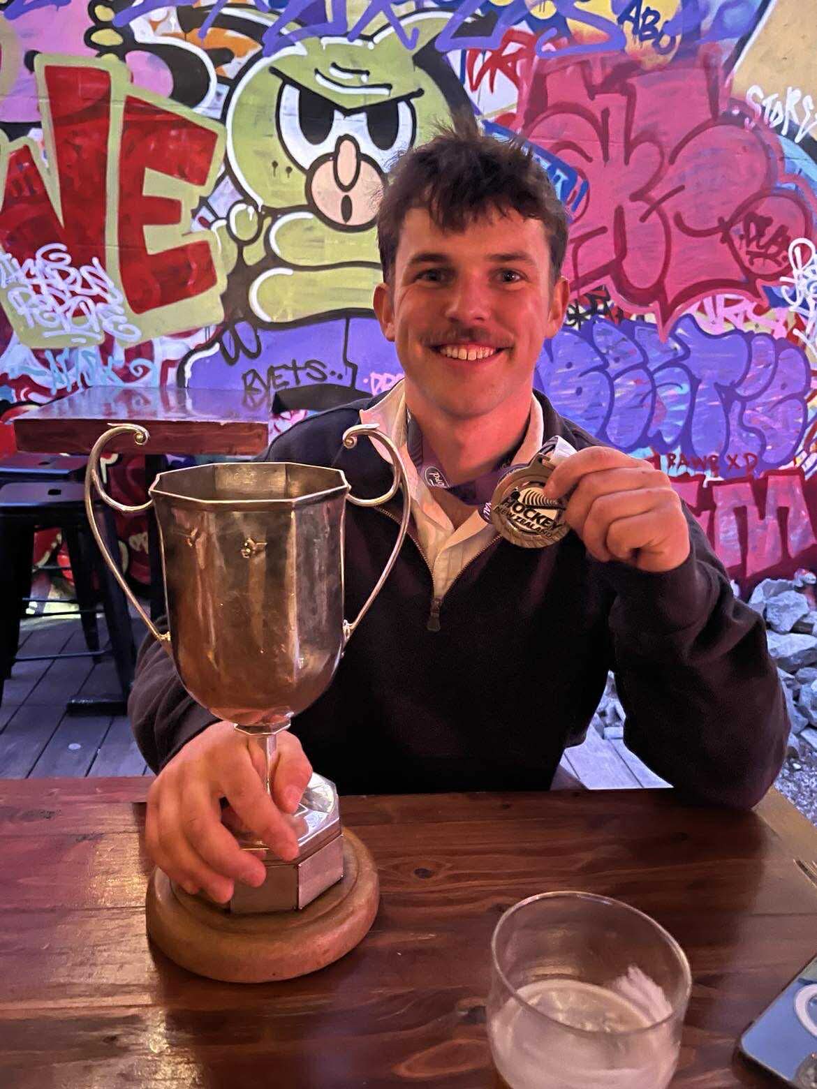 Smiling man with a mustache holding a large trophy and a medal in front of a colorful graffiti wall.