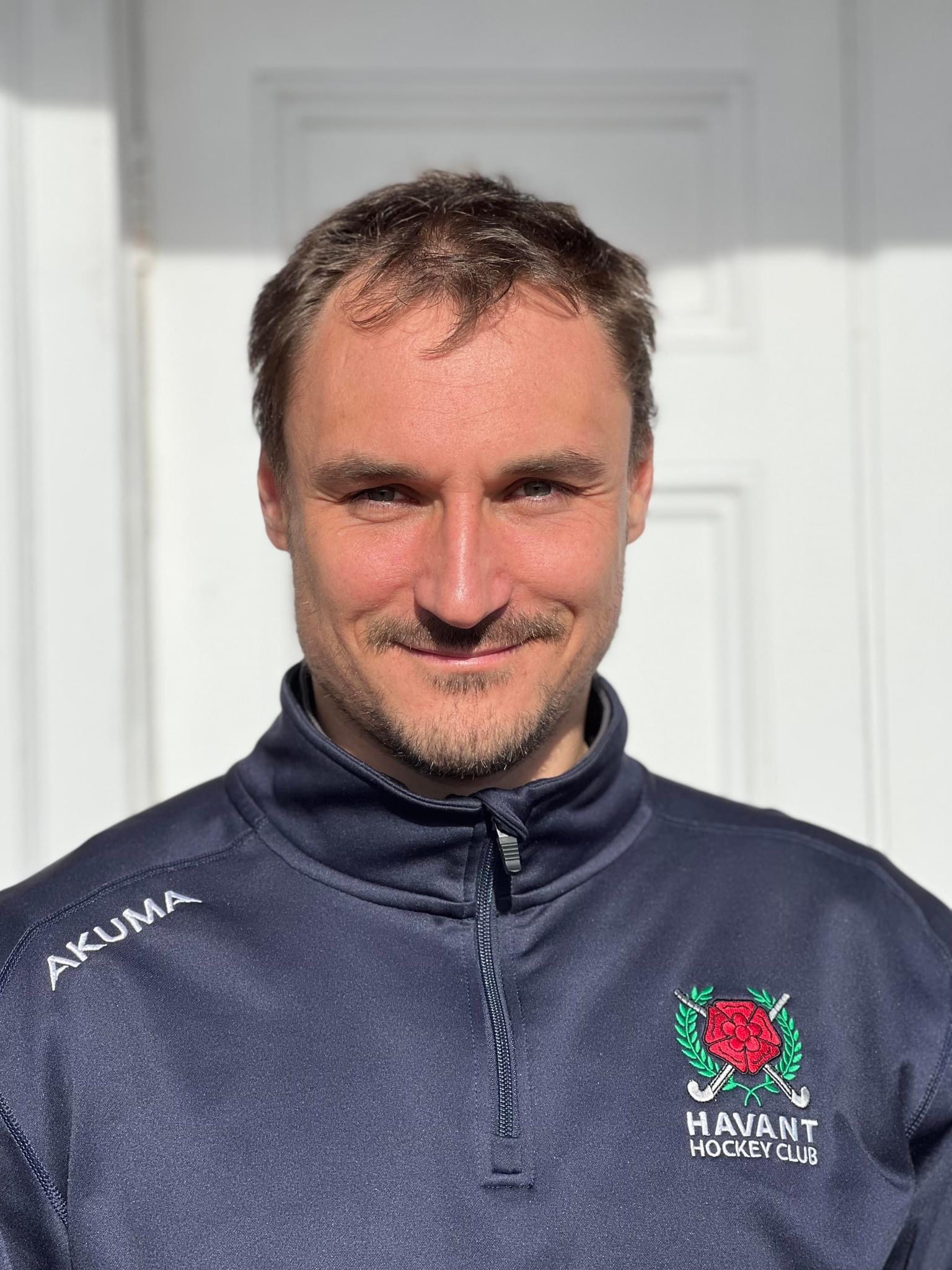 Man with short brown hair and light facial hair wearing a navy blue Havant Hockey Club zip-up jacket.