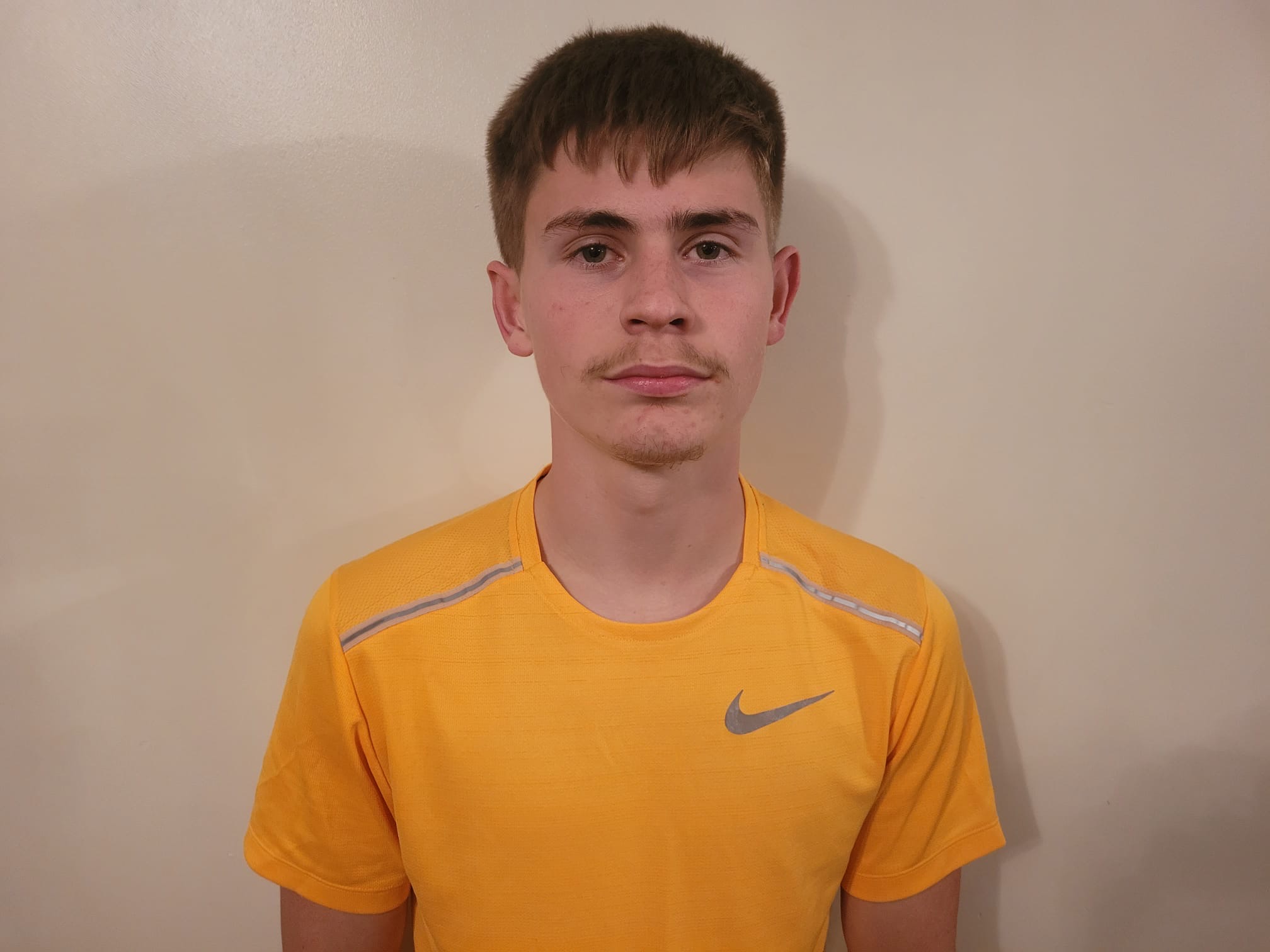 Young man with short brown hair and a faint mustache wearing a yellow Nike shirt against a plain beige wall.