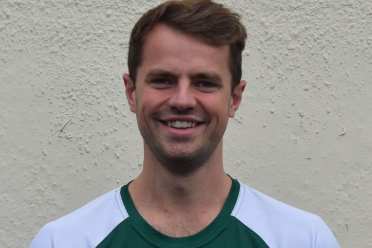 Smiling young man with short brown hair wearing a green and white shirt against a textured light wall.