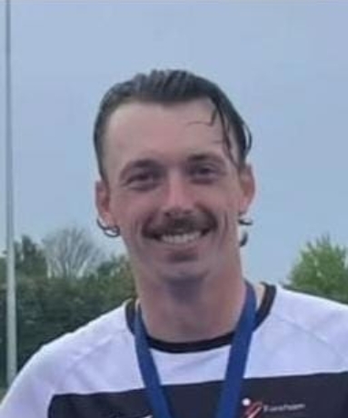 Smiling man with a mustache and short dark hair wearing a white and black striped shirt and a blue lanyard outdoors.