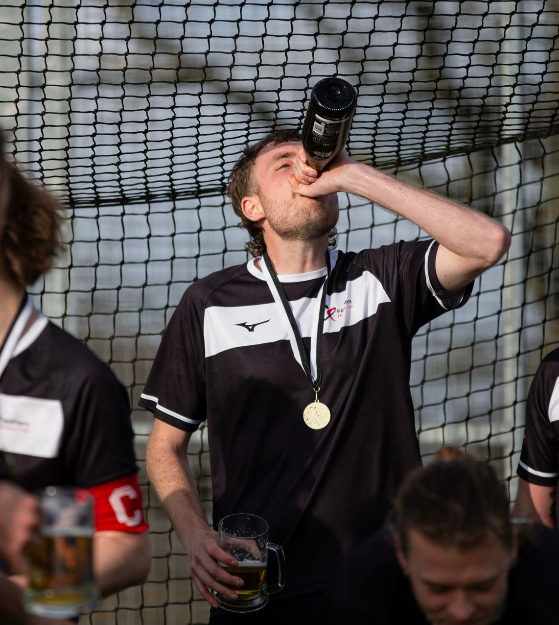Man wearing a sports jersey and a medal drinking from a bottle while holding a beer mug, standing in front of a net.