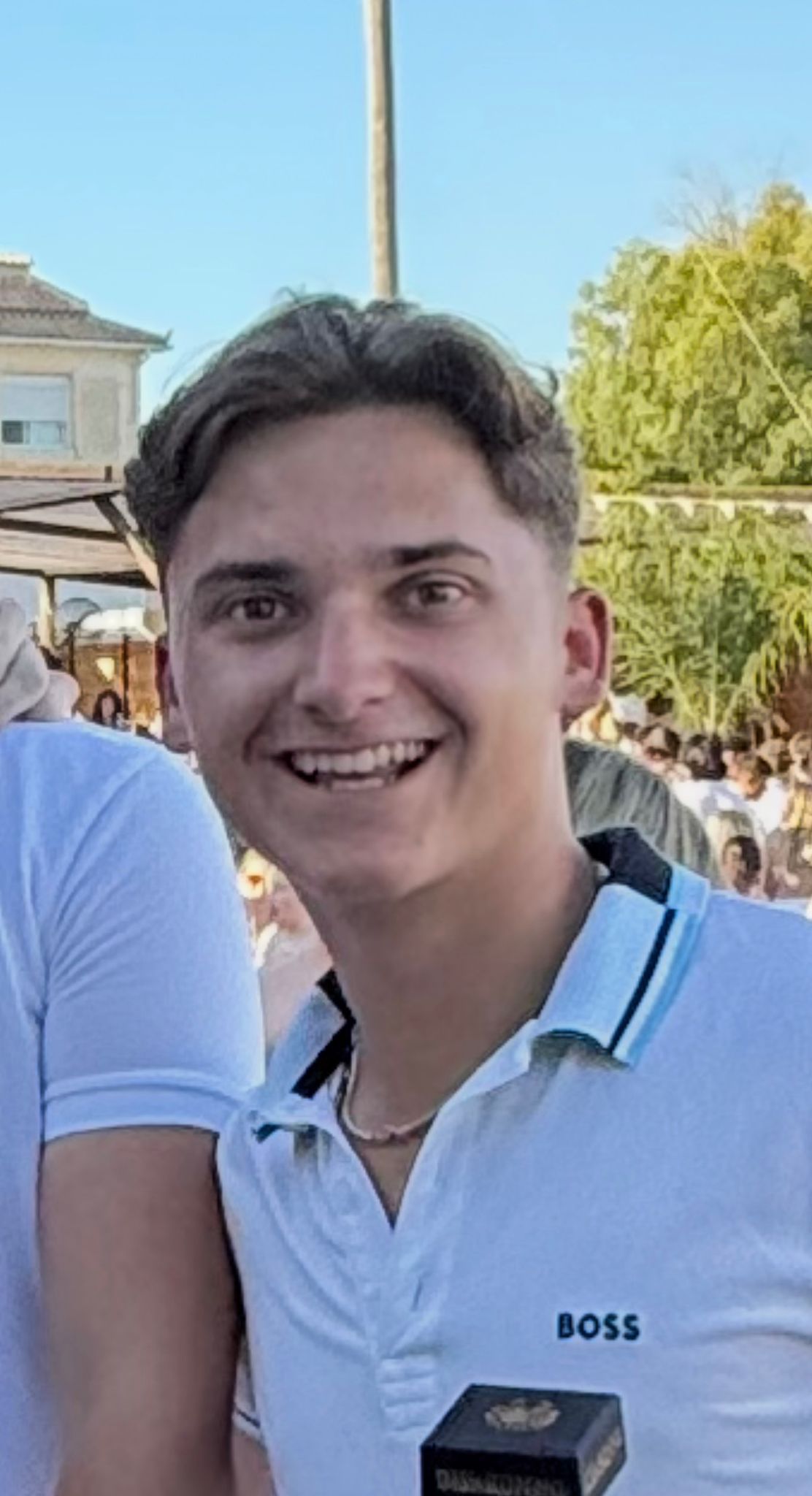 Smiling young man wearing a white BOSS polo shirt at an outdoor event on a sunny day.