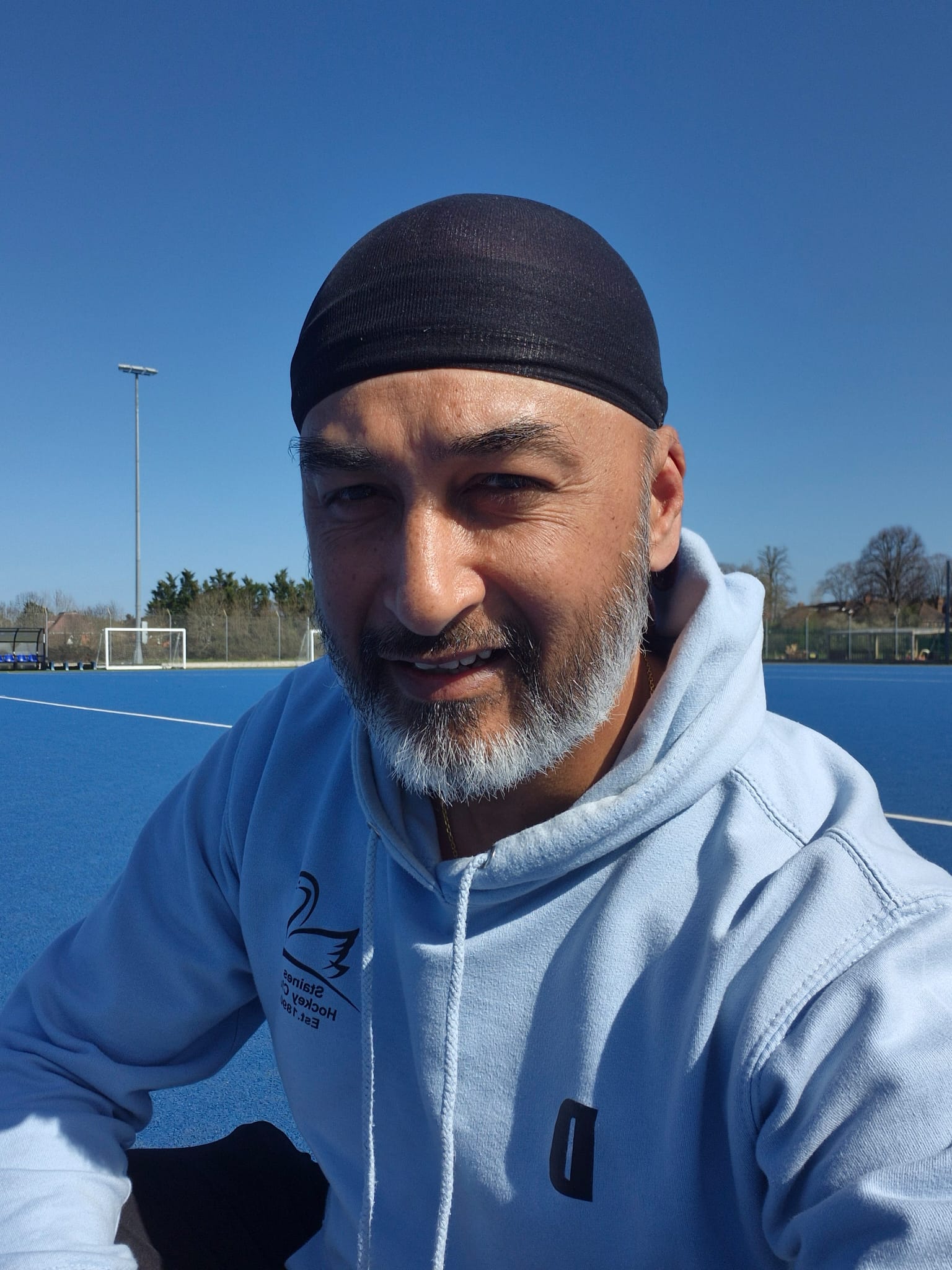 Man with a gray beard wearing a black head covering and light blue hoodie sitting on a blue sports field under a clear sky.