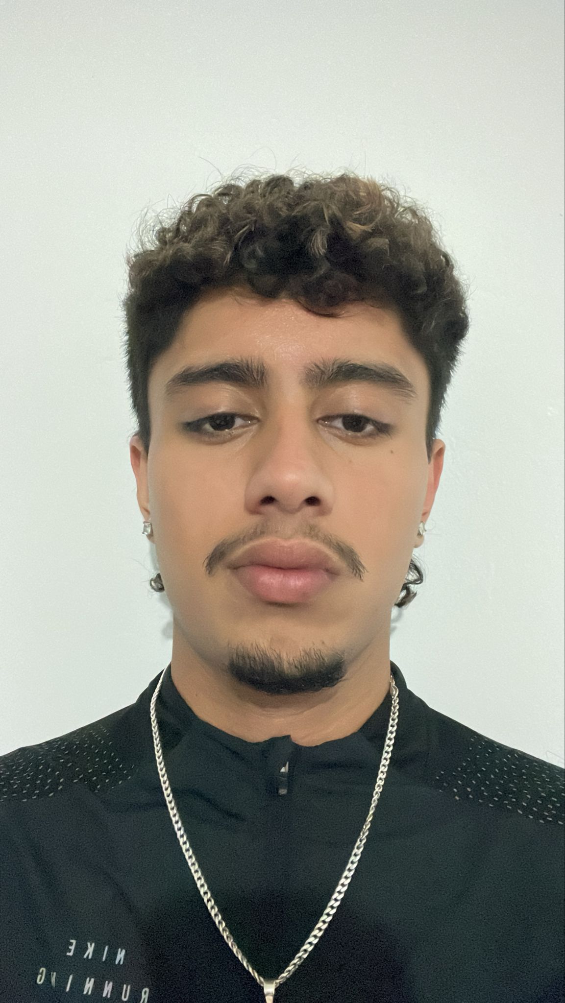 Young man with curly hair, light facial hair, wearing silver earrings, a silver chain necklace, and a black Nike running top.