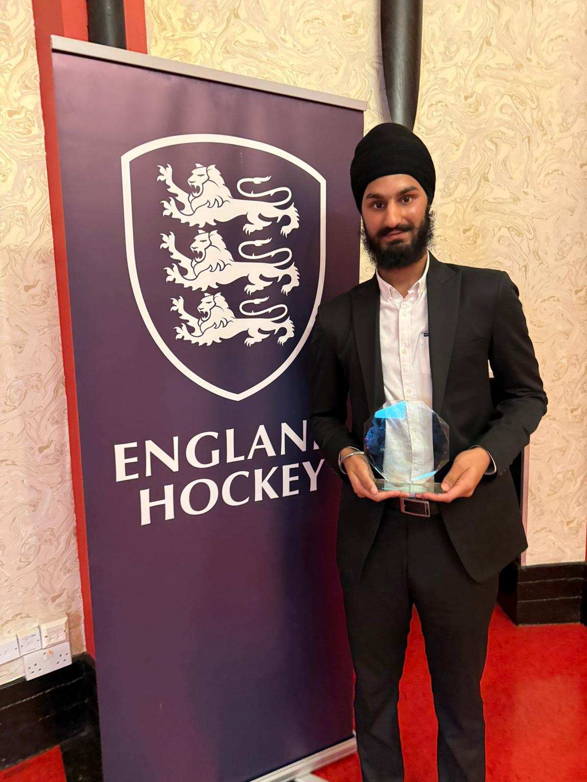 Man wearing a black turban and suit holding a glass award standing next to an England Hockey banner.
