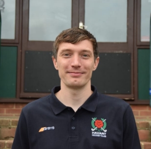 Young man with short brown hair wearing a black Havant Hockey Club polo shirt, standing outdoors in front of a building with brick and windows.