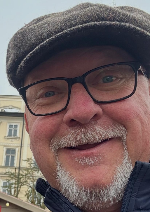 Close-up of a smiling middle-aged man wearing black glasses, a brown flat cap, and a dark jacket with light facial hair, standing outdoors with a building in the background.