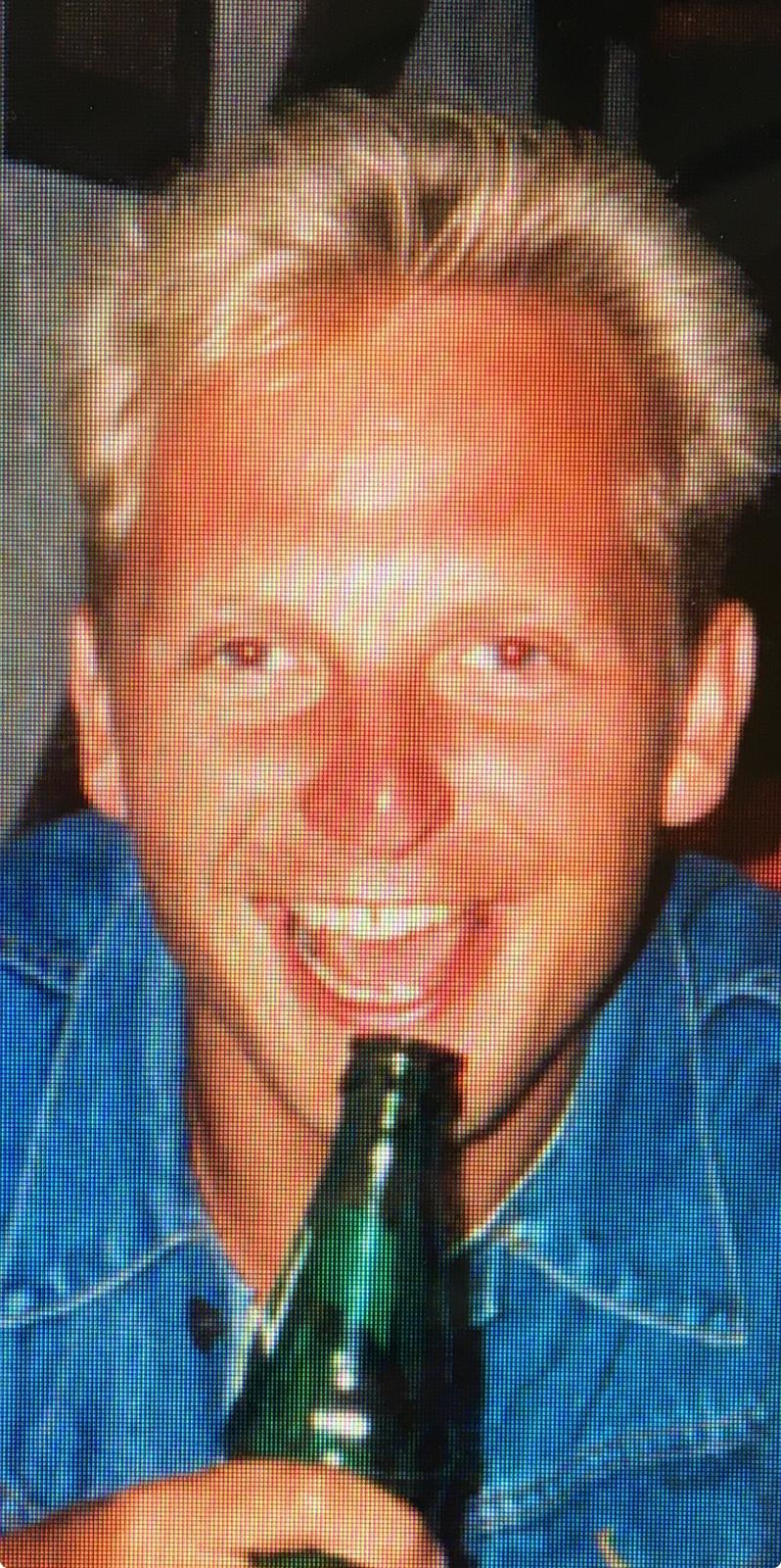 Smiling man with blonde hair wearing a blue denim shirt holding a green glass bottle near his mouth.