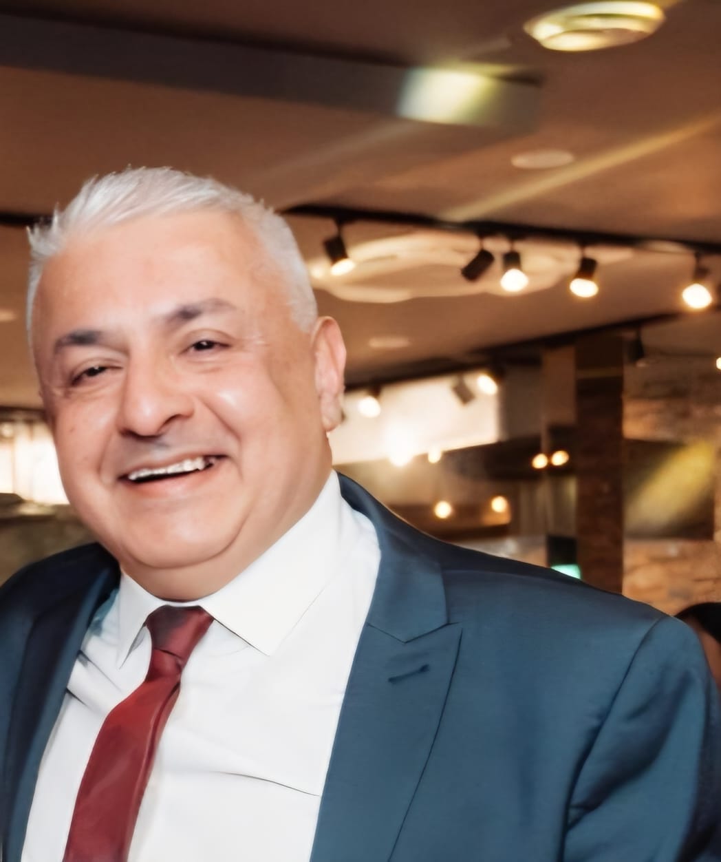 Smiling man with short white hair wearing a dark suit jacket, white shirt, and red tie in a warmly lit indoor setting.