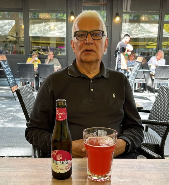 Elderly man with white hair and glasses sitting at an outdoor café table with a bottle and a glass of red Hoegaarden Rosée beer.
