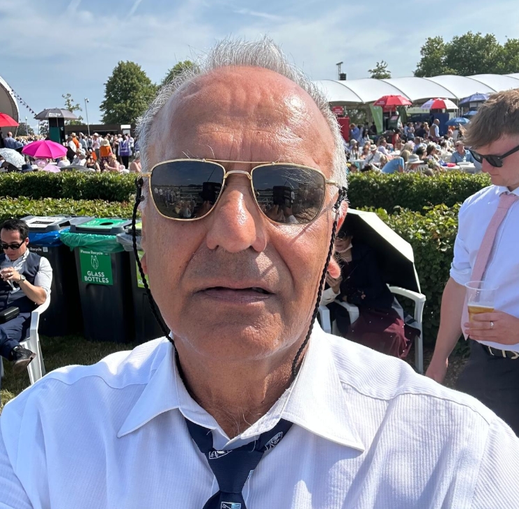 Older man wearing sunglasses, a white shirt, and a dark tie at a crowded outdoor event with people sitting and standing in the background.