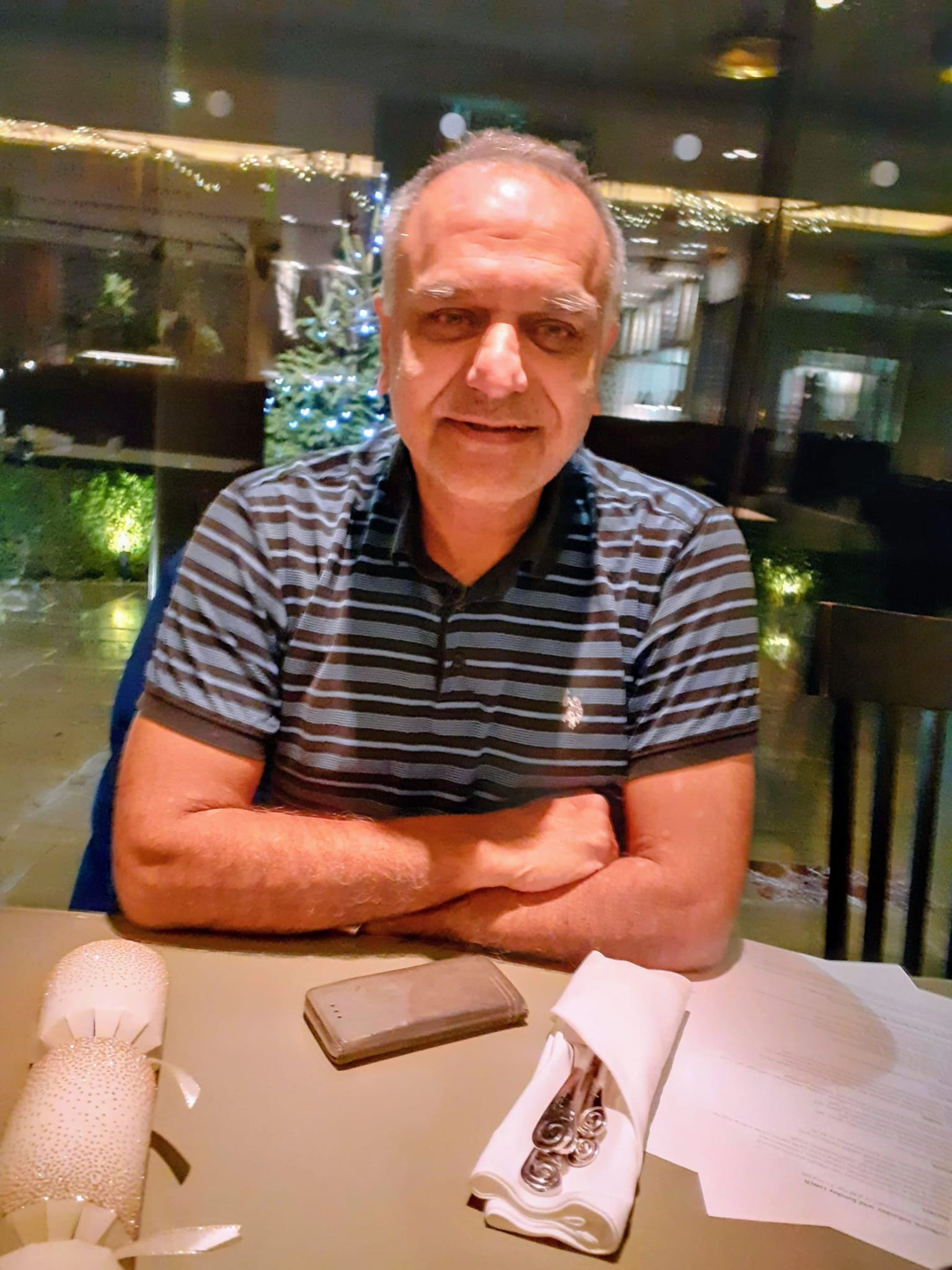 Man in a black and gray striped polo shirt sitting at a table with crossed arms, with tableware wrapped in a white napkin, a phone, and a menu on the table, and a decorated Christmas tree in the background.