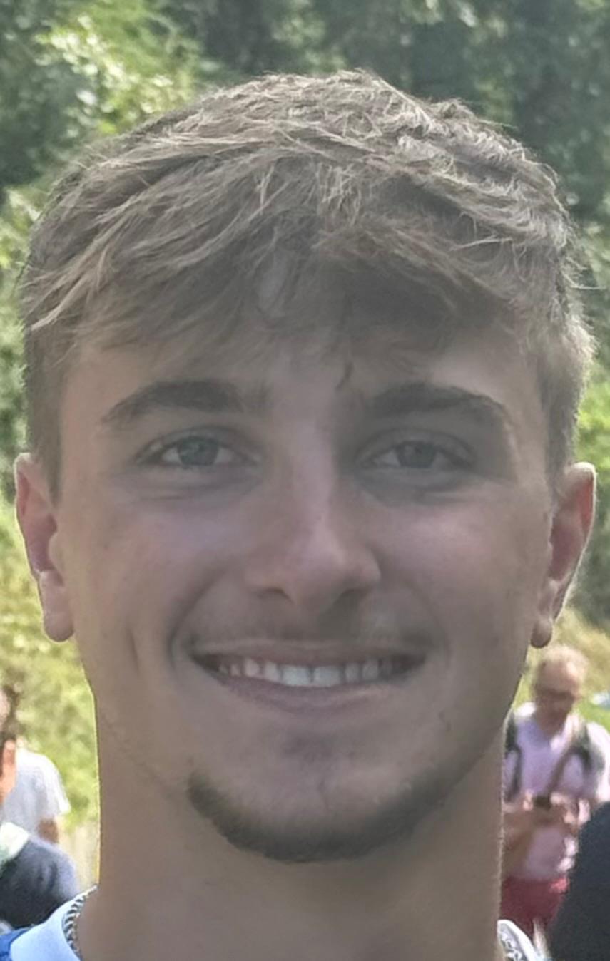 Close-up of a young man with light brown hair and a goatee smiling outdoors with trees and people in the blurred background.