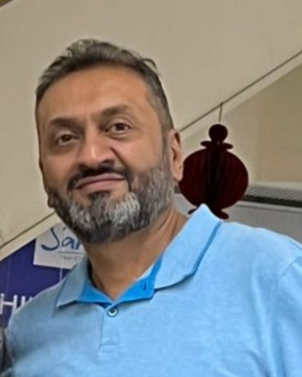 Bearded man wearing a light blue polo shirt standing indoors near a staircase with a decorative element behind him.