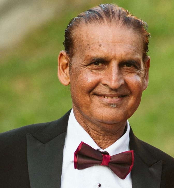 Smiling older man wearing a black tuxedo jacket, white shirt, and maroon bow tie with an outdoor green background.