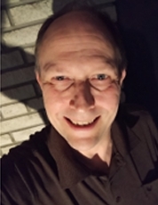 Smiling middle-aged man in a dark shirt standing in front of a brick wall at night.
