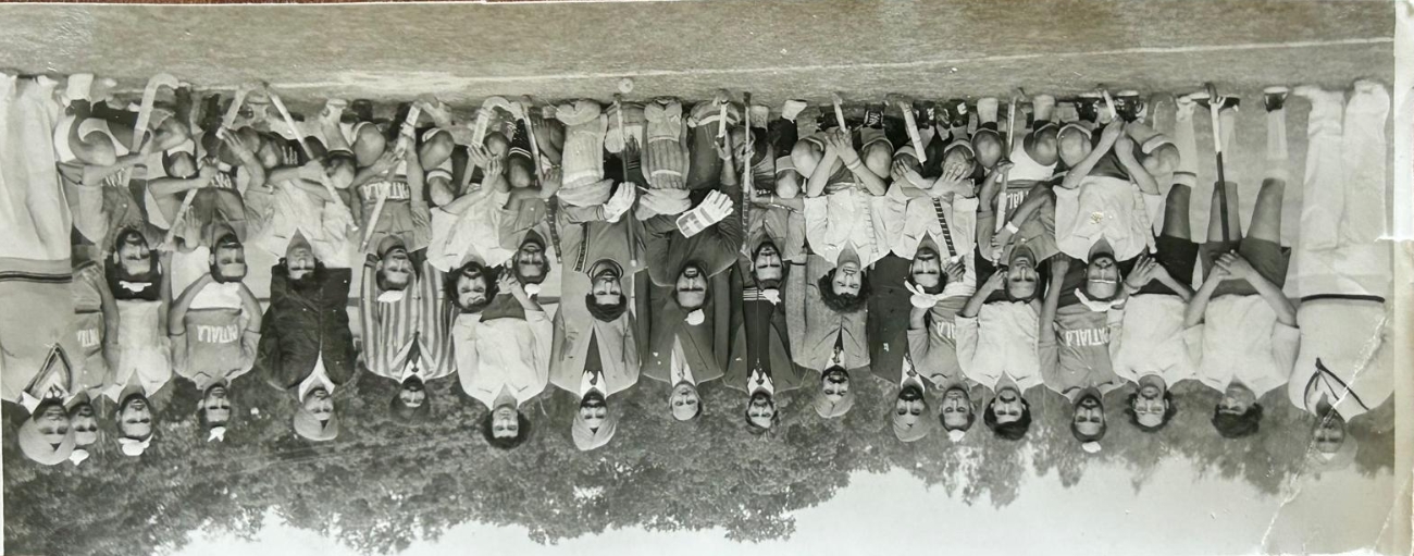 Group photo of a men's soccer team with 19 members on grass field, 16 wearing orange jerseys and white shorts, 3 in casual clothes, and one lying in front in a red shirt and black pants.