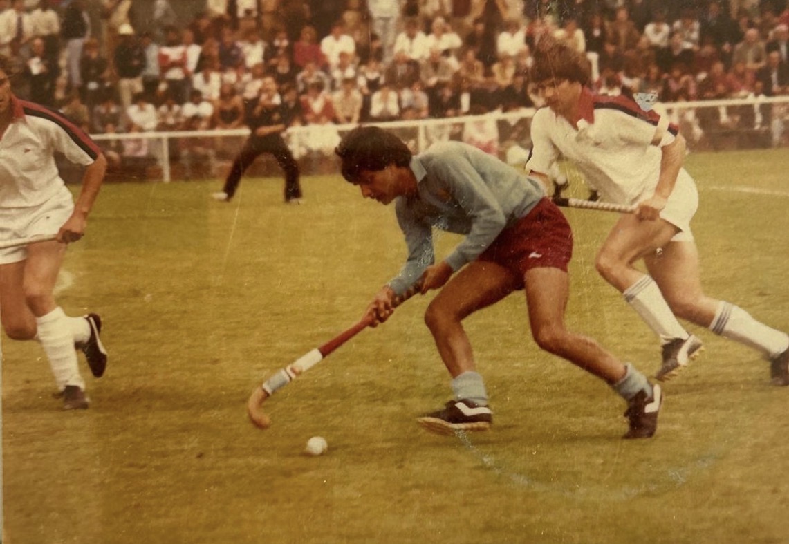 Group photo of a men's soccer team with 19 members on grass field, 16 wearing orange jerseys and white shorts, 3 in casual clothes, and one lying in front in a red shirt and black pants.
