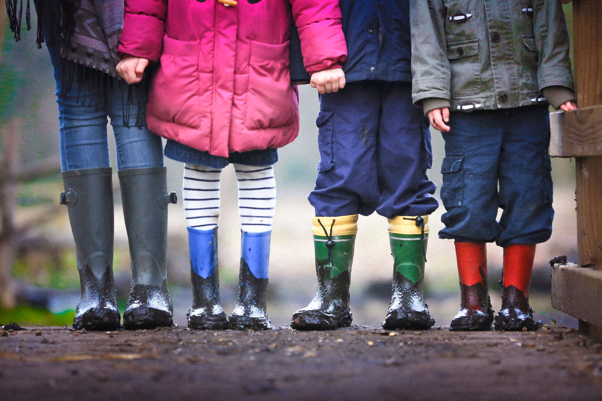 Four children standing side by side on a muddy path, wearing colorful rubber boots and holding hands, with their legs and boots covered in mud up to their ankles.