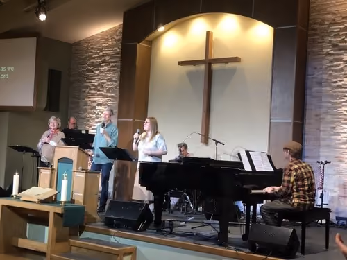A church worship band performing on stage with four singers and a pianist in front of a large wooden cross mounted on a wall under warm lighting.