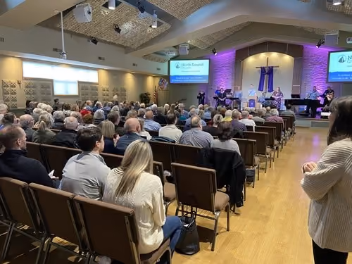 Congregation seated in rows of chairs inside a church facing the front where a worship band and speakers perform on a stage with purple lighting and cross backdrop.