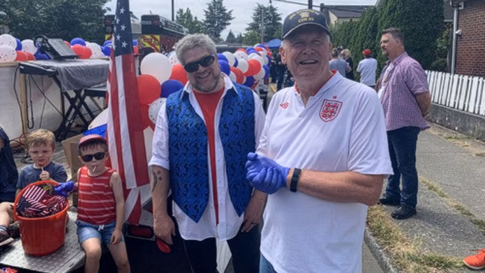 Two men smiling outdoors at a festive event with red, white, and blue balloons and an American flag; one man wears sunglasses, a blue vest, and a red shirt while the other wears a white shirt and a black cap. Two children sit nearby, one wearing sunglasses and a red striped tank top, the other holding a small American flag.