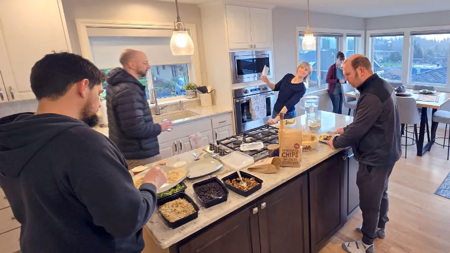 Four adults stand around a kitchen island in a modern open kitchen, with one person serving themselves food from trays and others holding cups, as a woman smiles and gestures towards the group.