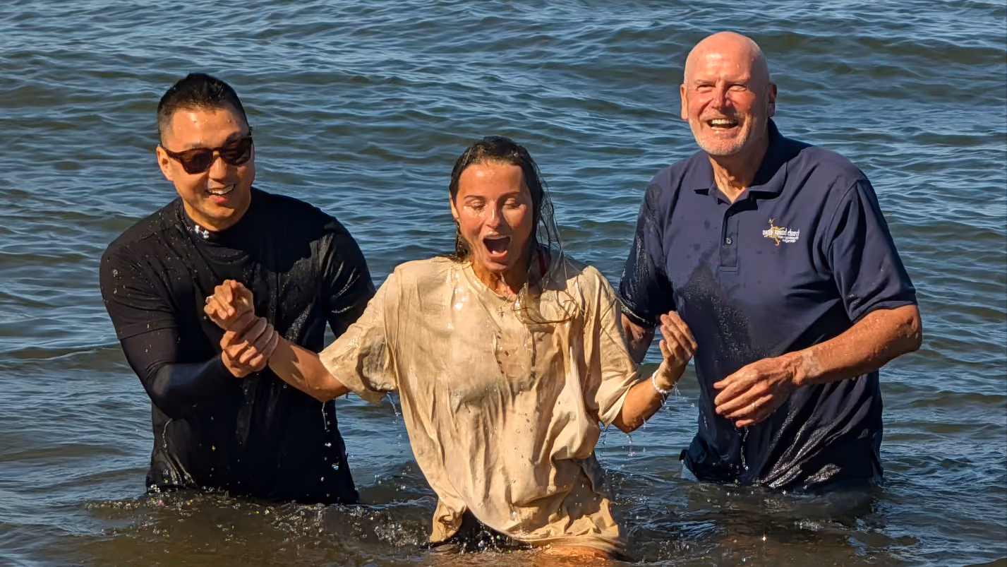 Three people waist-deep in water during a baptism ceremony: a woman in the center with wet hair and a soaked beige shirt, holding hands with two men on either side, one wearing sunglasses and a black shirt, the other bald with a blue polo shirt, both smiling.